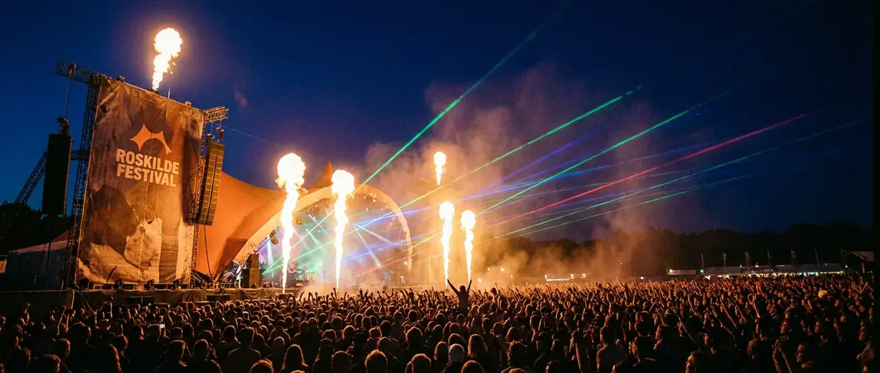 Publikum står tæt sammen foran en oplyst scene til en koncert.